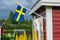 Swedish flag on traditional Swedish small red wooden guest cottage, white window, door. Summer day after rain, yellow flowers,