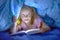 Sweet beautiful and pretty little blond child girl 6 to 8 years old lying under bed covers reading book in the dark with torch fla