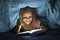 Sweet beautiful and pretty little blond girl 6 to 8 years old under bed covers reading book in the dark at night with torch light