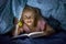 Sweet beautiful and pretty little blond girl 6 to 8 years old under bed covers reading book in the dark at night with torch light