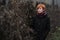 Sweet red-haired girl in a black coat and purple knitted scarf is standing by the fence overgrown with grapevine or ivy