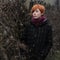 Sweet red-haired girl in a black coat and purple knitted scarf is standing by the fence overgrown with grapevine or ivy, girl or w