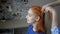 A sweet, smiling teenage girl is sitting in a room by the window, and her mother is braiding her red hair into pigtails