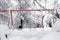 Swing for children in garden in winter, covered with snow, only silhouettes of red metal construction are visible.