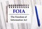 On the table are charts and reports, on which lie a blue pen and a notebook with the word FOIA The Freedom of Information Act