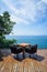 Table and rattan outdoor armchair setting on wooden deck and glass balcony with panoramic Andaman ocean sea view, green tree
