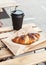 Take away cappuccino or black coffee in cup and fresh french chocolate croissant in paper bag on wooden terrace table