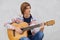 Talented teenage boy with trendy hairdo wearing shirt and jeans holding acoustic guitar playing his favourite songs while sitting