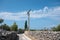 Tall concrete pole with electric wire in countryside. Narrow road with stone wall. Blue sky with white clouds.