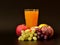 A tall, faceted glass of a mixture of fruit juices on a black background, next to a bunch of grapes, an apple, a peach, a