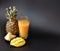 A tall faceted glass of a mixture of fruit juices on a black background, next to pieces of ripe pineapple and mango