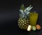 A tall faceted glass of a mixture of fruit juices on a black background, next to pieces of ripe pineapple and pieces of kiwi