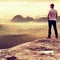 Tall man in white shirt and black trousers with red baseball cap stay on sharp cliff and watch to valley. Colorful misty mornin