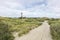 Tall red and white striped lighthouse building standing on small hill. Sandy coastal landscape with green vegetation and footpaths