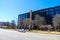 A tall rusty smoke stack in front of a black and glass office building surrounded yellow winter grass and bare winter trees