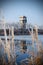 Tall watchtower reflection on icy water, viewed between frozen plants