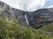 Tall waterfall at Romsdalen valley with rocks and green forest. Blue sky white clouds background. Norway summer landscape