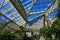 A tall white metal and glass greenhouse filled with lush green trees and plants and colorful flowers with blue sky