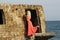 Tanned blonde girl in a red dress stands on the stone ruins of an old fort, against the backdrop of the waves of the Baltic Sea
