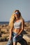 Tanned blonde girl in sunglasses and jeans sits on the rocks on the beach on a sunny day