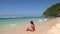 Tanned brunette woman in red swimsuit sitting near water, enjoying coming foam waves on wild tropic beach with blue