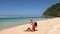 Tanned brunette woman in red swimsuit sitting near water, enjoying coming waves and pour coconut juice over on wild