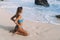 Tanned girl with sporty body in swimsuit sits on sandy beach and looks on ocean and waves