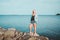 Tanned woman posing at the beach on summer day, looking up. Dreamy female in jeans clothes, standing rocky coast, blue