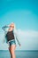 Tanned woman posing at the beach on summer day, looking up. Dreamy female in jeans clothes, standing rocky coast, blue