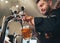 Tapping fresh lager beer in glass mug close up. Smiling stylish bearded barman dressed black uniform with an apron at bar counter