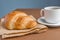 Tasty breackfast. French croissant served on craft paper and cup of black coffee or espresso on brown background
