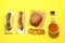Tasty burger, hot dogs, fried onion rings, different sauces and refreshing drink on yellow background, flat lay. Fast food