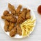 Tasty fried chicken drumsticks, spicy wings, French fries, chicken tenders and sauce on white plate over white wooden surface, top