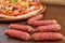 Tasty little sausages salami on brown wooden table close up. On blurred background pizza with tomatoes, bell pepper, onion