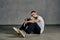 Tattooed fellow with beard. Dressed in white t-shirt, sneakers, black sports trousers. Sitting sideways on floor, gray