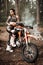 Tattooed racer girl wearing motocross outfit with semi naked torso posing next to her bikein the woods