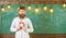 Teacher in eyeglasses holds alarm clock. Schedule and regime concept. Bearded hipster holds clock, chalkboard on