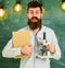 Teacher in eyeglasses holds book and microscope. Science concept. Scientist holds book and microscope, chalkboard on