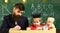 Teacher in formal wear and pupil in mortarboard in classroom, chalkboard on background. Father checking homework, helps