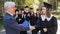 The teacher shakes hands with the student and presents the diploma outdoors. A group of university graduates.