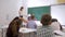 Teacher stands at front of class near blackboard and asks pupils question to which they raise their hands