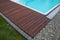 Teak wood pool deck detail next decorative stones and grass, natural hardwood material installed around the blue swimming pool