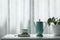 Teal cup and tall teapot with notebook and silver pen on white table with plant pot in front of a see-through curtain
