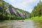 A team of people floats on a river in an inflatable catamaran among beautiful rocks and green coniferous forests with a clear blue