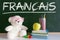 Teddy bear wearing glasses in a French language classroom next to an apple and a pile of books and a dictionary