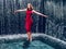 Teen ager girl in high fashion red dress posing in water, water fall behind her back. Prom photoshoot in beautiful location Young