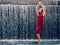 Teen ager girl in high fashion red dress posing in water, water fall behind her back. Prom photoshoot in beautiful location Young