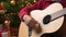 Teen boy playing guitar, sitting indoor near decorated xmas tree with lights, dressed as Santa helper - Merry Christmas and Happy