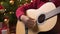 Teen boy playing guitar, sitting indoor near decorated xmas tree with lights, dressed as Santa helper - Merry Christmas and Happy