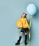 Teen child in yellow sweatshirt, black skirt, knee-highs, boots. Smiling, holding balloon and gift box, posing on blue background.